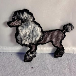 May include: An embroidered iron-on patch shaped like a poodle dog. The poodle features a textured, fluffy-looking grey and white head and chest, with a dark brown, grid-patterned body, legs, and tail, all outlined in a thick black border.