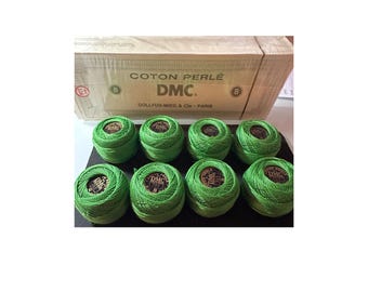 May include: Eight spools of bright green DMC Coton Perlé size 8 embroidery thread. The spools are arranged in two rows on a black surface, with a cardboard box in the background. The box is labeled "COTON PERLÉ DMC".