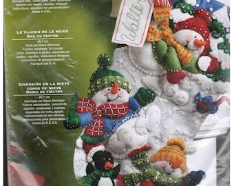 May include: A Bucilla Snow Fun felt stocking kit package featuring a white stocking decorated with three snowmen, a penguin, and a blue bird. The kit includes stamped felts, cotton floss, sequins, beads, needles, and instructions for an 18-inch stocking.