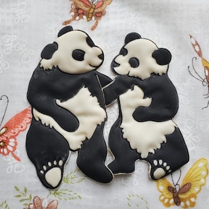 Puede incluir: Dos osos panda negros y blancos están cosidos sobre un fondo de tela blanca con un patrón floral. Los pandas se miran y parecen estar bailando.