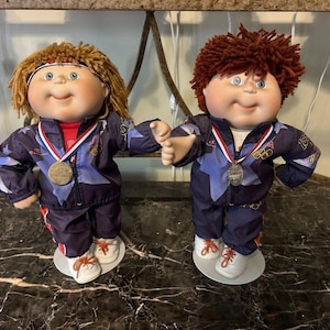 May include: Two Cabbage Patch Kids dolls wearing blue and red tracksuits with Olympic rings and medals. The dolls are standing on white bases.