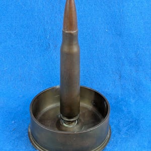 May include: A metallic candle holder featuring a central upright bullet-shaped component attached to a circular base. The object has a dark, aged bronze-toned finish with visible patina and a smooth, cylindrical structure throughout its design.