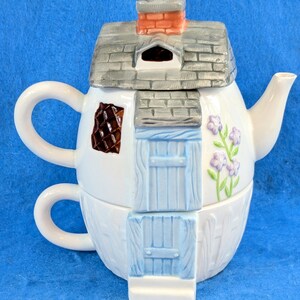 May include: A ceramic teapot and teacup stack designed to resemble a cottage. The white teapot features a gray shingled roof with a red brick chimney, a blue door, and a brown window. The white teacup base has a matching blue door and fence.