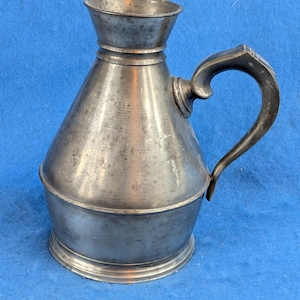 May include: A metallic pitcher with a wide, flared rim, a conical body, and a curved handle. The surface has a weathered, matte gray finish with visible signs of oxidation and wear, featuring a distinct horizontal ridge near the base and neck.