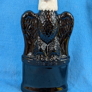 May include: A dark brown glass decanter shaped like an eagle with its wings spread. The eagle has a white head and a textured feather pattern on its body. The base of the decanter features the embossed text "1776-1976" along the front rim.