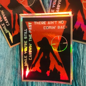 May include: A rectangular holographic sticker featuring a black silhouette of a person against a red and orange sunset background. The text reads, "WHILE YOU'RE STILL CARRYIN' THE PAST" and "THERE AIN'T NO COMIN' BACK," with a crosshair symbol.