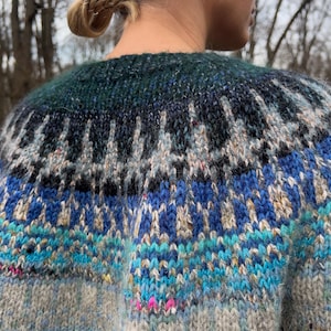 May include: A close-up of a knitted jumper with a colourful design. The jumper features a pattern of blue, teal, black, and white geometric shapes. The top portion is a dark teal colour, and the bottom is a light grey.