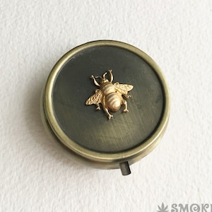 May include: A small, round, brass trinket box with a hinged lid. The lid features a gold-colored bee design.