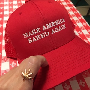 May include: Red baseball cap with a white mesh back. The cap has white embroidered text that reads "Make America Baked Again".