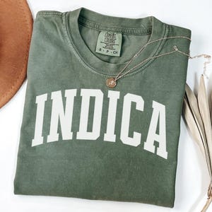 May include: An olive green t-shirt with the word "INDICA" printed in large white block letters. The shirt has a crew neck and a small tag at the neckline. A gold necklace with a round pendant rests on the shirt.
