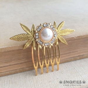 Peut inclure: Un peigne à cheveux doré avec une perle centrale entourée de cristaux transparents. Le peigne présente un motif floral avec des accents de feuilles et quatre dents. La marque "SMOKIES" est visible en bas.