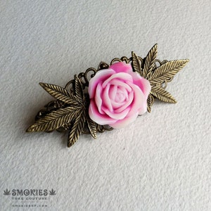 Peut inclure: Barrette décorative avec une rose rose et blanche au centre, entourée de feuilles et de filigranes de couleur bronze. La barrette porte le texte "SMOKIES" et "TOKE COUTURE" en bas.
