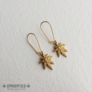 May include: A pair of gold dangle earrings featuring a marijuana leaf design. The earrings are made of metal and have a simple, minimalist style.
