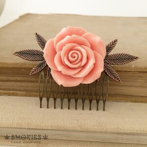 Peut inclure: Un peigne à cheveux avec une rose pêche et des feuilles et dents en bronze. La rose est le point central, avec des pétales détaillés. Le peigne est posé sur un fond neutre, soulignant le motif floral. Le texte "SMOKIES TOKE COUTURE" est en bas.
