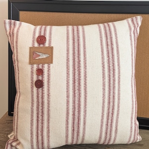 May include: A white and red striped throw pillow with three red buttons and a small brown fabric patch with a design on it.