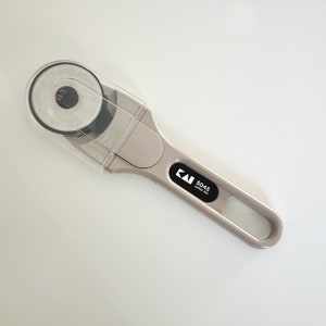 Może przedstawiać: Beigeowy obcinak rotacyjny z przezroczystą plastikową osłoną i czarną rączką. Na rączce nadrukowany jest napis "KAI 5045 JAPAN PKT".