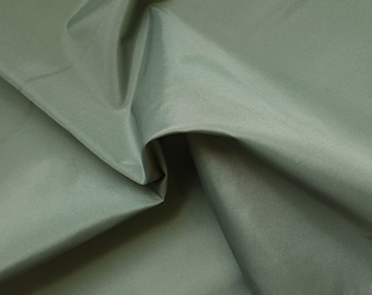 May include: A solid sage green fabric with a tight, uniform weave and a smooth, slightly matte finish. The material is draped to create soft folds and deep shadows, highlighting the consistent texture and muted green color of the textile.