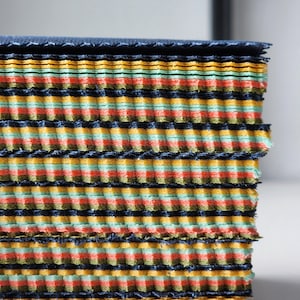 May include: A stack of rectangular fabric swatches featuring a repeating horizontal pattern of dark navy blue, mustard yellow, light mint green, and coral orange stripes. The edges of the fabric layers are textured with a distinct corrugated finish.