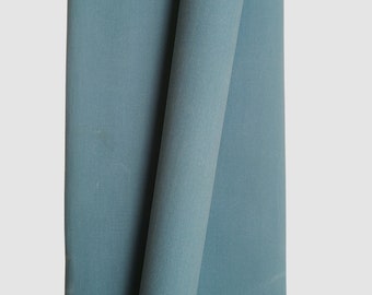 May include: A close-up of a folded, teal-colored fabric. The fabric is a solid color and appears to be a medium-weight material. The fabric is folded over itself, creating a layered effect. The background is a plain, light gray.