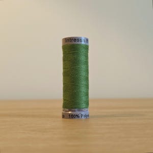 Puede incluir: Un carrete de hilo de coser color verde oliva con forma cilíndrica. La etiqueta superior dice Intressa y la inferior dice 100% Polyester. El hilo está enrollado firmemente alrededor del núcleo central, creando una textura suave.