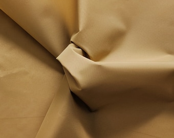 May include: A solid tan fabric with a smooth, matte texture. The material is draped to create soft folds and deep shadows, highlighting the consistent color and fine weave of the textile surface across the entire visible area of the cloth.