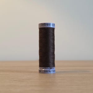 Könnte beinhalten: Eine zylindrische Spule mit dunkelbraunem Nähgarn und weißen Etiketten an der Ober- und Unterseite. Das obere Etikett trägt die Aufschrift Intressa, das untere 100% Polyester. Der Faden ist fest um den Kunststoffkern gewickelt.