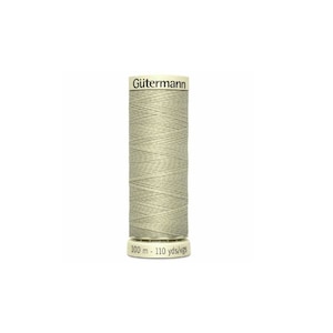 Può includere: Un'bobina di filo da cucire Gütermann di colore beige chiaro. La bobina è etichettata "Gütermann" e "100 m - 110 yds/vgs".