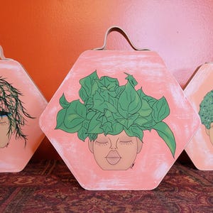May include: Three hexagonal wall art pieces with a peach background and a painted face with closed eyes. Each piece features a different hairstyle: green leaves, green textured hair, and green spiky hair. They have brown leather handles.