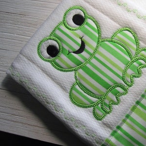 Puede incluir: Un babero blanco para bebé con una aplicación de rana de rayas verdes y blancas. La rana tiene ojos negros y una sonrisa negra.