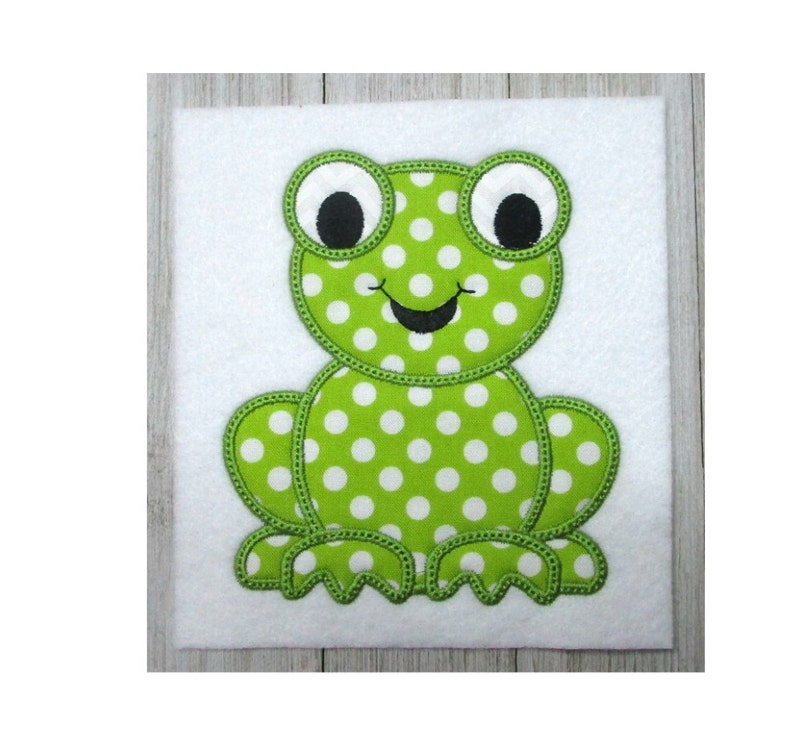 May include: Green felt applique of a frog with white polka dots. The frog has large black eyes and a stitched-on smile.