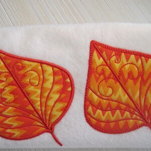 K&ouml;nnte beinhalten: Zwei applizierte Blattformen in Orange- und Gelbt&ouml;nen mit roter Umrandung und dekorativen N&auml;hten. Die Bl&auml;tter sind vor einem wei&szlig;en Hintergrund platziert und schaffen ein herbstliches Design.