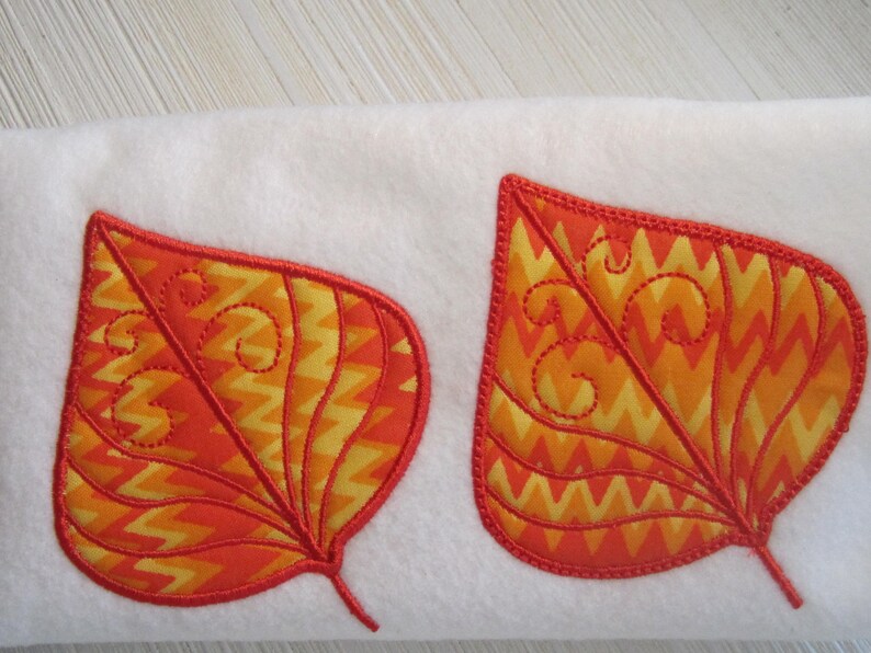 K&ouml;nnte beinhalten: Zwei applizierte Blattformen in Orange- und Gelbt&ouml;nen mit roter Umrandung und dekorativen N&auml;hten. Die Bl&auml;tter sind vor einem wei&szlig;en Hintergrund platziert und schaffen ein herbstliches Design.