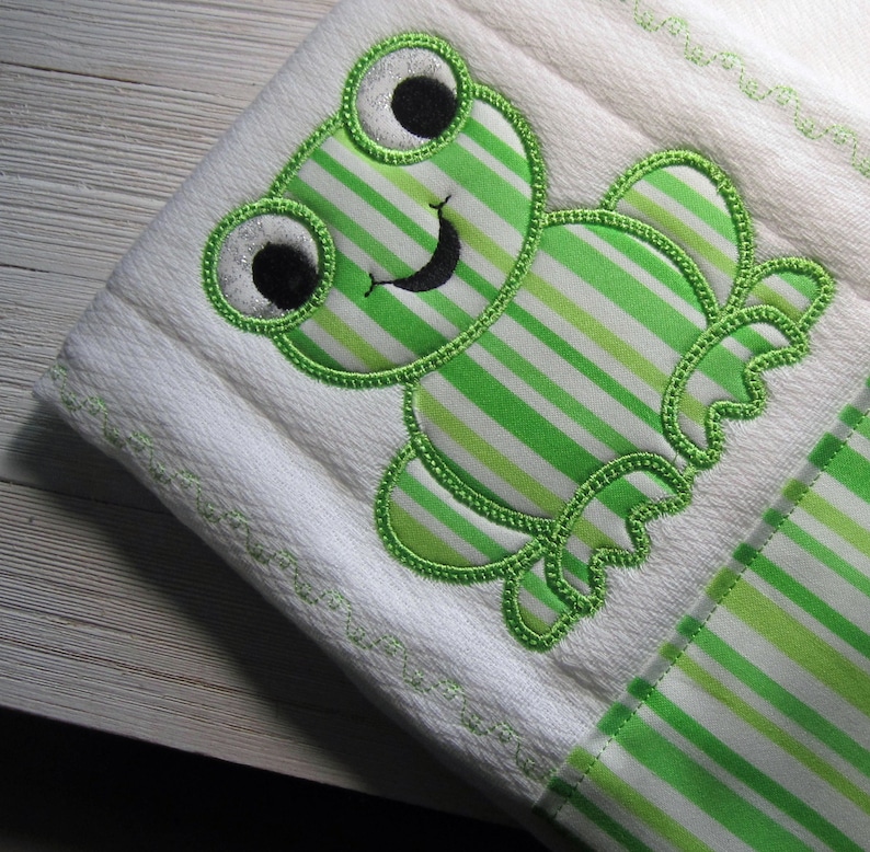 May include: A white baby burp cloth with a green and white striped frog applique. The frog has black eyes and a black smile.