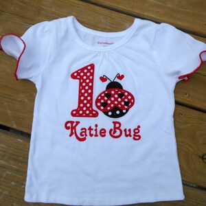 May include: White t-shirt with red and black ladybug and the number 1 with red polka dots. The text "Katie Bug" is embroidered in red below the ladybug.