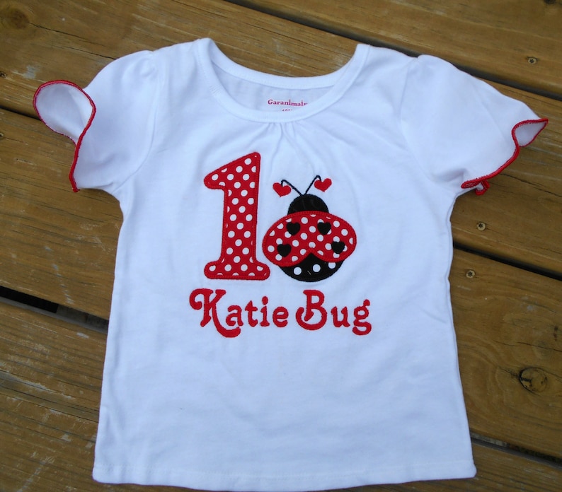 May include: White t-shirt with red and black ladybug and the number 1 with red polka dots. The text "Katie Bug" is embroidered in red below the ladybug.