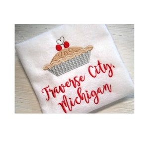 Puede incluir: Toalla blanca bordada con un diseño de pastel de cerezas rojas y el texto "Traverse City, Michigan".