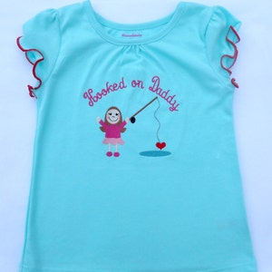 May include: A light blue t-shirt with a pink embroidered design of a girl holding a fishing rod with a heart on the end. The text "Hooked on Daddy" is embroidered in pink on the shirt.
