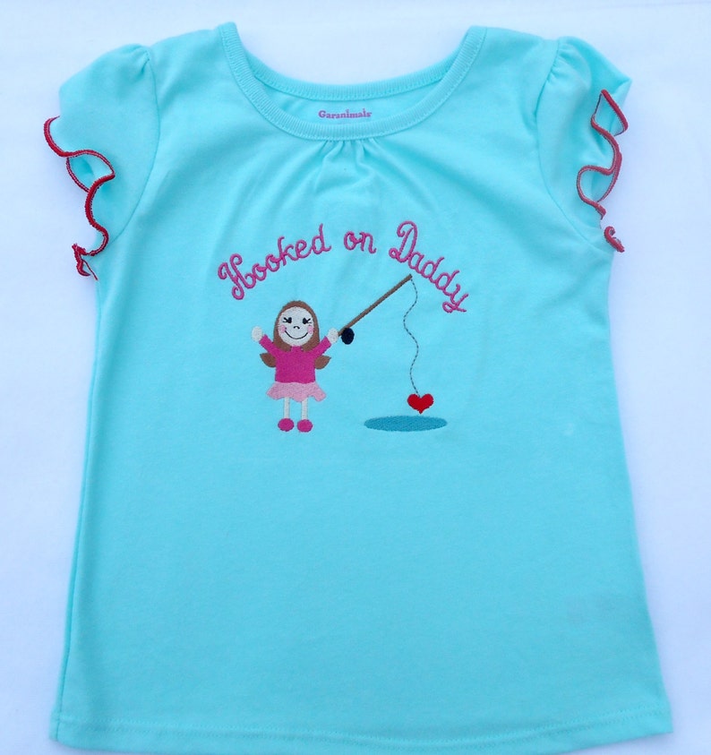 May include: A light blue t-shirt with a pink embroidered design of a girl holding a fishing rod with a heart on the end. The text "Hooked on Daddy" is embroidered in pink on the shirt.