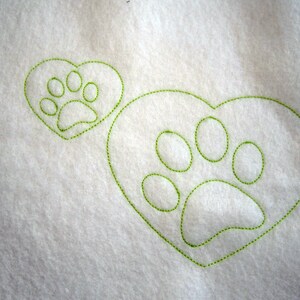 May include: A white fabric background with two green embroidered paw prints. One paw print is inside a heart shape and the other is a larger paw print.