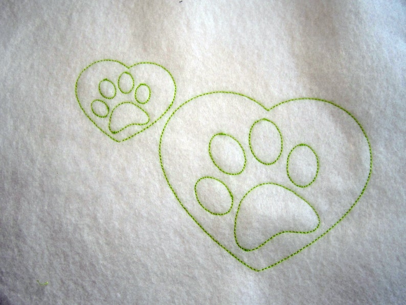May include: A white fabric background with two green embroidered paw prints. One paw print is inside a heart shape and the other is a larger paw print.