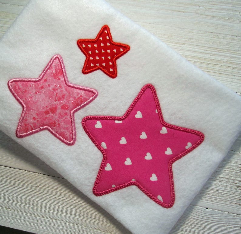 May include: Three embroidered stars on a white fabric background. The largest star is pink with white hearts, the medium star is pink with white hearts, and the smallest star is red with white hearts.