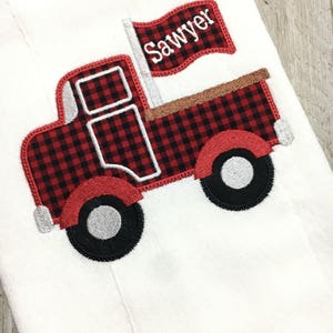May include: White fabric featuring an embroidered red and black check truck design. The truck has black wheels with silver centres and a flag that reads "Sawyer". The truck bed is brown.
