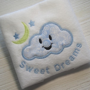 Puede incluir: Manta de beb&eacute; blanca con bordado azul y verde de una nube sonriente, una luna creciente y estrellas. El texto "Sweet Dreams" est&aacute; bordado debajo de la nube.