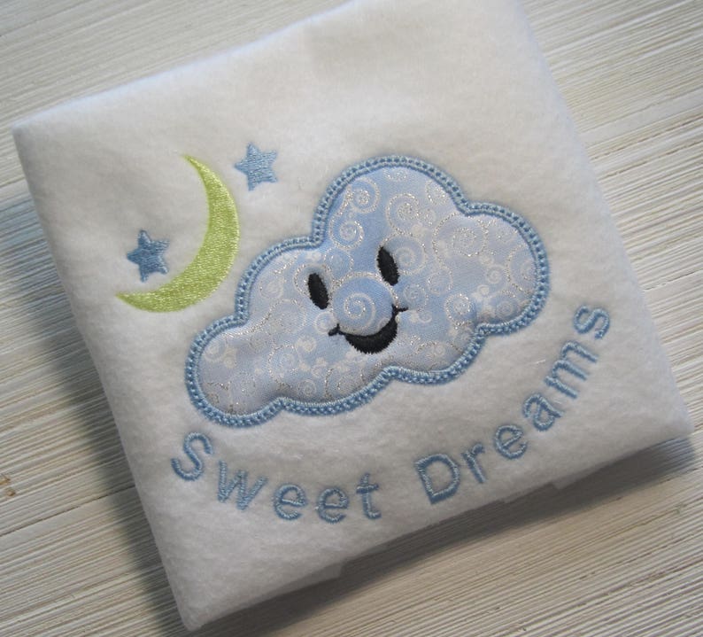Puede incluir: Manta de beb&eacute; blanca con bordado azul y verde de una nube sonriente, una luna creciente y estrellas. El texto "Sweet Dreams" est&aacute; bordado debajo de la nube.