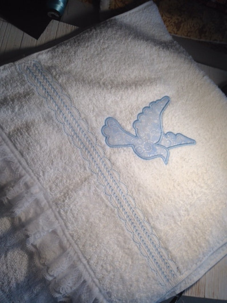 May include: White bath towel with a blue embroidered dove design and a white lace trim.