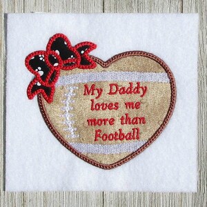 May include: Embroidered heart-shaped patch with a football design. The patch is brown with white stitching and a white stripe. It says "My Daddy loves me more than Football". A red bow with white polka dots is on top of the heart.