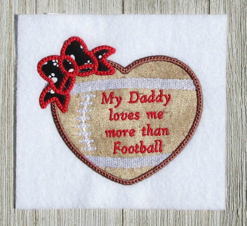 May include: Embroidered heart-shaped patch with a football design. The patch is brown with white stitching and a white stripe. It says "My Daddy loves me more than Football". A red bow with white polka dots is on top of the heart.