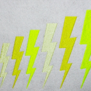 May include: A series of embroidered lightning bolts in varying shades of yellow and chartreuse are arranged diagonally on a light gray background. The bolts increase in size from left to right, creating a visual gradient.
