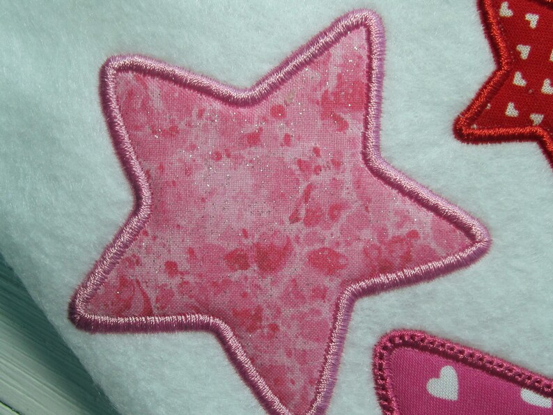 May include: A pink star with a glittery finish and a pink stitched border. The star is on a white background.