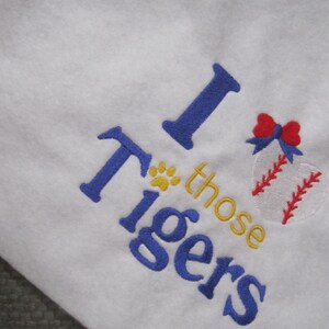 May include: White fabric with blue embroidered text that reads "I those Tigers". A baseball with red stitching and a red bow is embroidered on the right side. A yellow paw print is above the word "those".